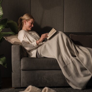 Eine junge Frau sitzt gemütlich und zugedeckt auf der Couch und liest ein Buch | © Karin Pasterer
